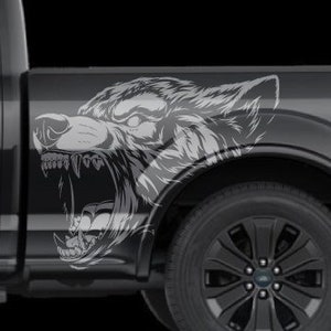 May include: A gray pickup truck with a wolf graphic decal on the side of the truck bed. The wolf is facing forward with its mouth open and teeth showing. The truck has black wheels and a black bumper.