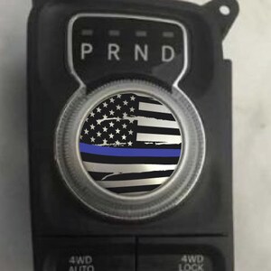 May include: A black and white American flag with a blue stripe across the middle. The flag is on a round, silver-colored knob with the letters "PRND" above it.