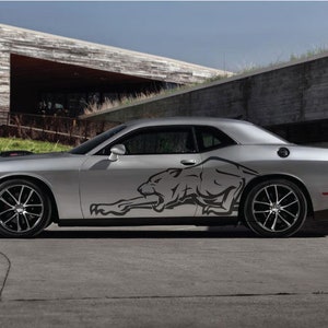 Op de afbeelding: Een zilveren Dodge Challenger met een zwarte panter grafische sticker aan de zijkant. De auto staat geparkeerd voor een modern gebouw met een stenen gevel.