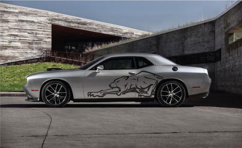Op de afbeelding: Een zilveren Dodge Challenger met een zwarte panter grafische sticker aan de zijkant. De auto staat geparkeerd voor een modern gebouw met een stenen gevel.