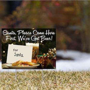 Puede incluir: Un cartel blanco con el texto "Santa, Please Come Here First, We've Got Beer!" y una nota que dice "for Santa." El cartel está delante de un vaso de cerveza, galletas y regalos envueltos.