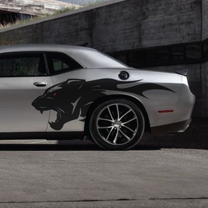 May include: A silver car featuring a black panther decal on its side. The panther has red eyes and a snarling expression. The car has black wheels and is parked on a concrete surface.