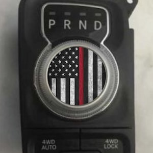 May include: Black vehicle gear shift panel with a circular emblem featuring a distressed American flag design. The flag includes white stars, white and red stripes, and a red vertical line. The panel also has the letters "P R N D" and buttons labeled "4WD AUTO" and "4WD LOCK."