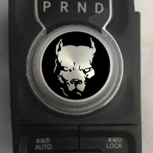 May include: A black and silver car gear shift knob with a black and white image of a pit bull dog's head. The gear shift knob has the letters "PRND" printed on it. The knob also has buttons labeled "4WD AUTO" and "4WD LOCK".