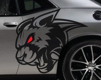以下が含まれることがあります： 銀色の車の側面に、赤い目をした黒と灰色の狼のステッカーが貼られています。ステッカーは車の後部座席側のドアとフェンダーに貼られています。