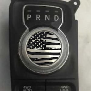 May include: A black and white American flag decal for a car gear shifter knob. The decal is in a distressed style with a silver metallic finish. The decal is placed on a black gear shifter knob with the letters "PRND" printed on it.
