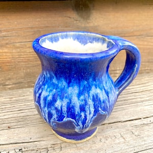 Könnte beinhalten: Eine handgefertigte Keramik-Tasse mit einer leuchtend blauen Glasur. Die Tasse hat einen abgerundeten Körper, einen stabilen Henkel und einen weißen Innenraum. Die blaue Glasur hat ein fließendes, strukturiertes Aussehen und erzeugt einen einzigartigen, künstlerischen Look.