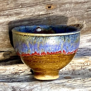 Könnte beinhalten: Eine kleine, braune Keramikschale mit einer blauen und violetten Glasur. Die Schale hat einen leicht ausgestellten Rand und einen abgerundeten Boden.