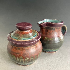 Könnte beinhalten: Ein Set aus zwei Keramik-Küchenutensilien, eine Zuckerdose mit Deckel und ein Milchkännchen. Beide Teile sind mit einer marmorierten Glasur in Braun-, Grün- und Lilatönen verziert.