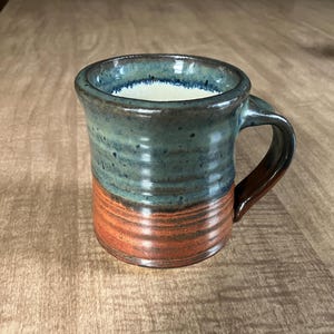 Könnte beinhalten: Eine handgefertigte Keramiktasse mit einer teal- und braunen Glasur. Die Tasse hat einen abgerundeten Körper und einen Henkel.