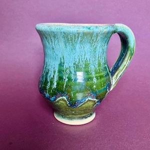 Könnte beinhalten: Eine handgefertigte Keramiktasse mit einer einzigartigen Glasur. Die Tasse hat einen grünen Boden, der oben in ein Türkisblau übergeht, mit einem Tropfglasureffekt. Der Griff ist gebogen und die Tasse hat eine weite Öffnung.