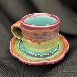 Könnte beinhalten: Ein farbenfrohes Keramik-Teetassen- und Untertassen-Set mit tealblauem Innenraum und mehrfarbiger Glasur. Die Tasse hat einen Henkel und die Untertasse einen gewellten Rand.