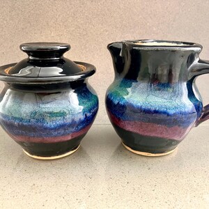 Könnte beinhalten: Ein Set aus zwei Keramik-Küchenutensilien in schwarzer, blauer und violetter Glasur. Die Teile sind ein Topf mit Deckel und eine Kanne.
