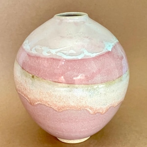 May include: A spherical ceramic vase with a narrow opening, featuring a layered glaze in shades of soft pink, cream, and white. The surface has a textured, matte finish with visible horizontal bands of colour and subtle drips of white glaze.