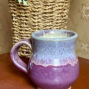May include: A ceramic mug with a rounded body and a curved handle. The mug features a two-tone glaze with a deep purple base that transitions into a lighter, mottled lavender and blue upper section, finished with a smooth, glossy texture.
