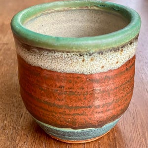 Könnte beinhalten: Eine handgefertigte Keramiktasse mit grünem Rand, einem weißen und beigen Band und einem rotbraunen Körper. Die Tasse hat eine abgerundete Form und eine strukturierte Oberfläche, ideal für Ihr Lieblingsgetränk.