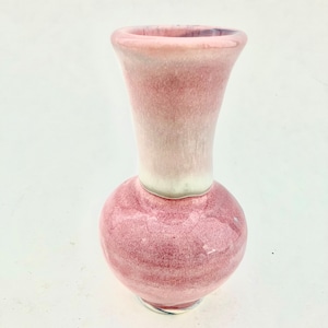 Könnte beinhalten: Eine kleine, rosafarbene Keramikvase mit abgerundetem Boden und einem ausgestellten Hals. Die Vase hat eine weiße Glasur auf der oberen Hälfte und eine rosafarbene Glasur auf der unteren Hälfte.