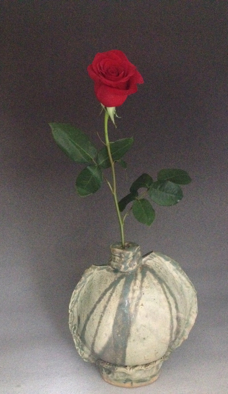 K&ouml;nnte beinhalten: Eine einzelne rote Rose mit gr&uuml;nen Bl&auml;ttern in einer wei&szlig;en und gr&uuml;nen Keramikvase mit strukturierter Oberfl&auml;che.