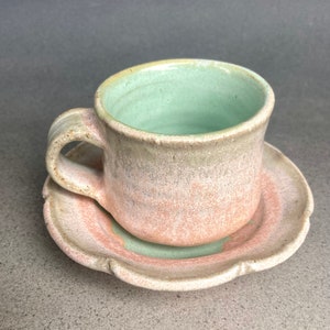 Könnte beinhalten: Ein hellrosa und grünes Keramik-Tee-Set aus Tasse und Untertasse. Die Tasse hat ein hellgrünes Inneres und die Untertasse einen gewellten Rand.