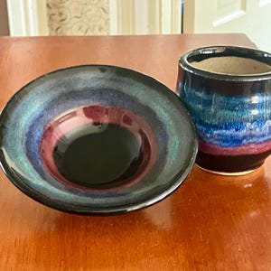 Könnte beinhalten: Ein Keramikschalen- und Becher-Set. Die Schale hat einen dunklen, glänzenden Innenraum mit konzentrischen Farbkreisen, darunter Blau, Lila und Schwarz. Der Becher hat ein ähnliches Farbschema mit Bändern in Blau, Türkis und Lila.