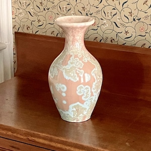 Könnte beinhalten: Eine dekorative Keramikvase mit einem zartrosa Sockel und einem ausgestellten Rand. Die Vase hat ein Muster aus weißen und hellblauen floralen Designs. Die Vase steht auf einer Holzoberfläche.