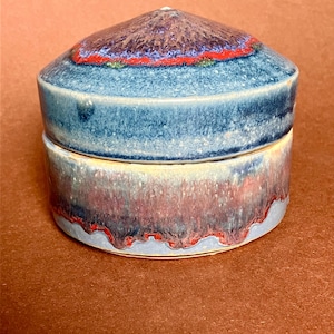 Può includere: Un vaso di ceramica con coperchio. Il vaso è cilindrico con un coperchio arrotondato. Il coperchio è viola scuro con una fascia rossa e blu. Il corpo del vaso è blu e rosso con un bordo rosso ondulato. Il vaso è su una superficie marrone.