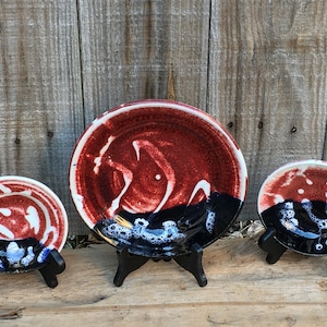 Könnte beinhalten: Drei Keramikteller mit roter, weißer und schwarzer Glasur. Die Teller werden auf schwarzen Ständern präsentiert.