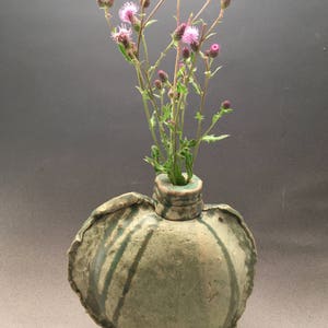 K&ouml;nnte beinhalten: Eine gr&uuml;ne Keramikvase mit einer einzigartigen, unregelm&auml;&szlig;igen Form und einer strukturierten Oberfl&auml;che. Die Vase ist mit einem Strau&szlig; rosa Wildblumen gef&uuml;llt.