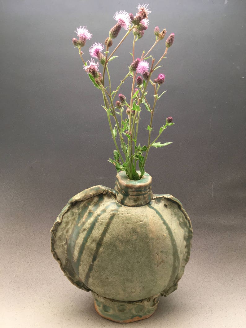 K&ouml;nnte beinhalten: Eine gr&uuml;ne Keramikvase mit einer einzigartigen, unregelm&auml;&szlig;igen Form und einer strukturierten Oberfl&auml;che. Die Vase ist mit einem Strau&szlig; rosa Wildblumen gef&uuml;llt.