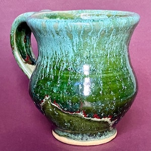 Könnte beinhalten: Eine handgefertigte Keramiktasse mit grüner und türkisfarbener Glasur. Die Tasse hat einen abgerundeten Körper, einen stabilen Henkel und einen leicht ausgestellten Rand. Die Glasur hat einen Tropfeffekt, mit roten und weißen Akzenten in der Nähe des Bodens. Die Tasse steht auf einem kleinen, unglasierten Sockel.