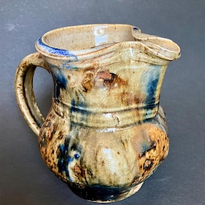 May include: A ceramic pitcher with a rounded body and a wide spout. The surface features a mottled glaze in shades of tan, brown, and deep blue, with a prominent horizontal ridge encircling the midsection and a curved handle on the side.