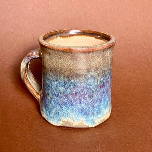 Könnte beinhalten: Eine handgefertigte Keramik-Tasse mit einer einzigartigen Glasur. Die Tasse weist einen Farbverlauf auf, der von Braun oben nach Blau unten übergeht. Der Griff und der Rand haben ein rustikales, strukturiertes Aussehen, ideal für Kaffee oder Tee.