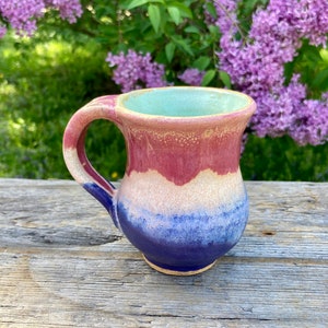 Könnte beinhalten: Eine handgefertigte Keramiktasse mit einer blauen, weißen und rosafarbenen Glasur. Die Tasse hat einen abgerundeten Körper und einen Henkel.