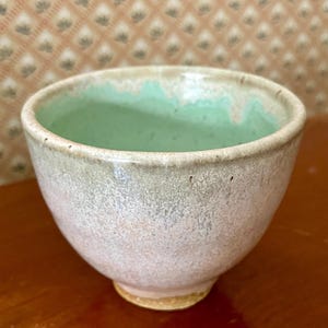 Könnte beinhalten: Eine kleine, handgefertigte Keramikschale mit runder Form. Die Außenseite ist in einem gesprenkelten, cremefarbenen Farbton gehalten, während die Innenseite eine hellgrüne Glasur aufweist. Die Schale steht auf einem kleinen, unglasierten Sockel.