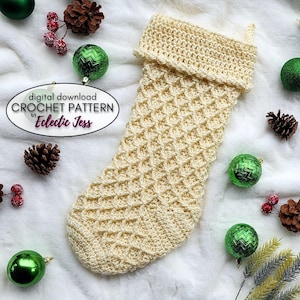 May include: A crocheted Christmas stocking in a cream colour with a diamond pattern. The stocking is on a white fluffy surface with green and red ornaments.