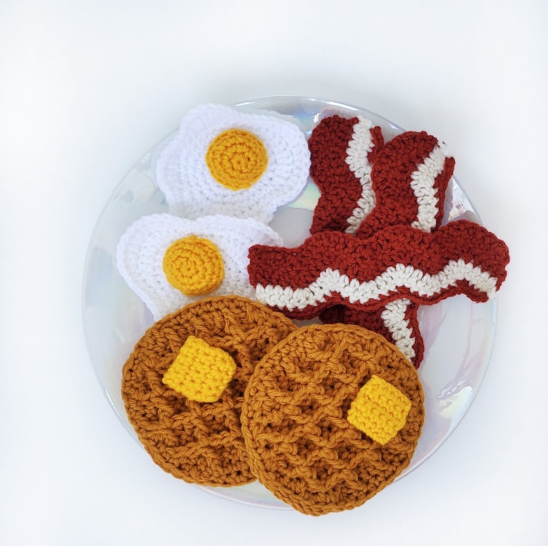 K&ouml;nnte beinhalten: H&auml;kelset mit Fr&uuml;hst&uuml;cksessen mit zwei gelben und wei&szlig;en Eiern, zwei braunen Waffeln und einer roten und wei&szlig;en Speckstreifen.