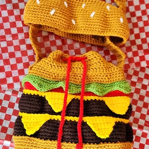 May include: A crocheted hamburger-shaped bag with a red drawstring and a matching crocheted bun hat. The bag is made with brown, yellow, green, and red yarn.