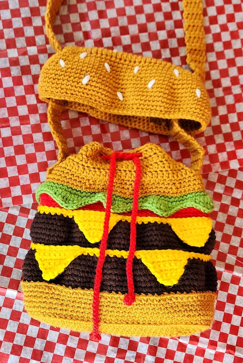 May include: A crocheted hamburger-shaped bag with a red drawstring and a matching crocheted bun hat. The bag is made with brown, yellow, green, and red yarn.