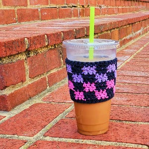 May include: A clear plastic cup with a crocheted pink and purple cup cozy. The cup is filled with iced coffee and has a green straw.