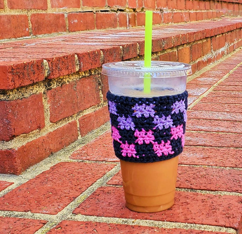 May include: A clear plastic cup with a crocheted pink and purple cup cozy. The cup is filled with iced coffee and has a green straw.