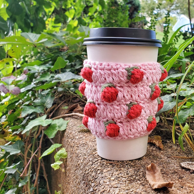 May include: A pink crocheted coffee cup cozy with red strawberry embellishments.
