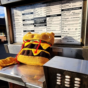 May include: A crocheted hamburger purse with a brown patty, yellow cheese, green lettuce, and red tomato. The purse is sitting on a metal surface with a call button.