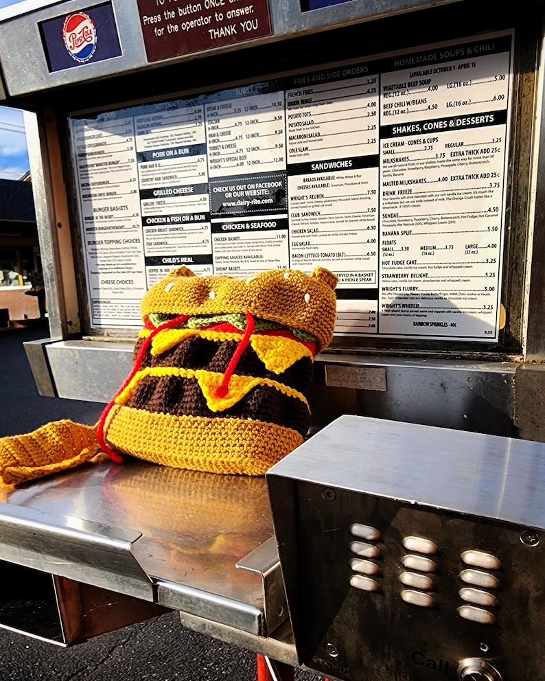 May include: A crocheted hamburger purse with a brown patty, yellow cheese, green lettuce, and red tomato. The purse is sitting on a metal surface with a call button.