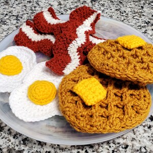 K&ouml;nnte beinhalten: Ein geh&auml;keltes Fr&uuml;hst&uuml;cksset auf einem wei&szlig;en Teller. Das Set enth&auml;lt zwei Eier, zwei Waffeln und drei Speckscheiben. Die Eier sind wei&szlig; mit gelben Dottern, die Waffeln sind braun und der Speck ist rot und wei&szlig;.