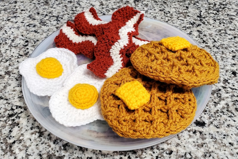 K&ouml;nnte beinhalten: Ein geh&auml;keltes Fr&uuml;hst&uuml;cksset auf einem wei&szlig;en Teller. Das Set enth&auml;lt zwei Eier, zwei Waffeln und drei Speckscheiben. Die Eier sind wei&szlig; mit gelben Dottern, die Waffeln sind braun und der Speck ist rot und wei&szlig;.