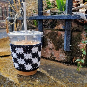 May include: A clear plastic cup with a black and white crocheted cup cozy. The cup is filled with a light brown beverage and has a white straw sticking out of the top.