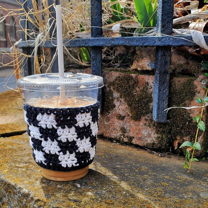 May include: A clear plastic cup with a black and white crocheted cup cozy. The cup is filled with a light brown beverage and has a white straw sticking out of the top.