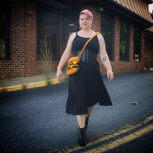 May include: A woman wearing a black tank top, a black corset, and a black pleated skirt. She is carrying a brown and yellow bag with a strap over her shoulder. She is wearing black boots and has pink hair.