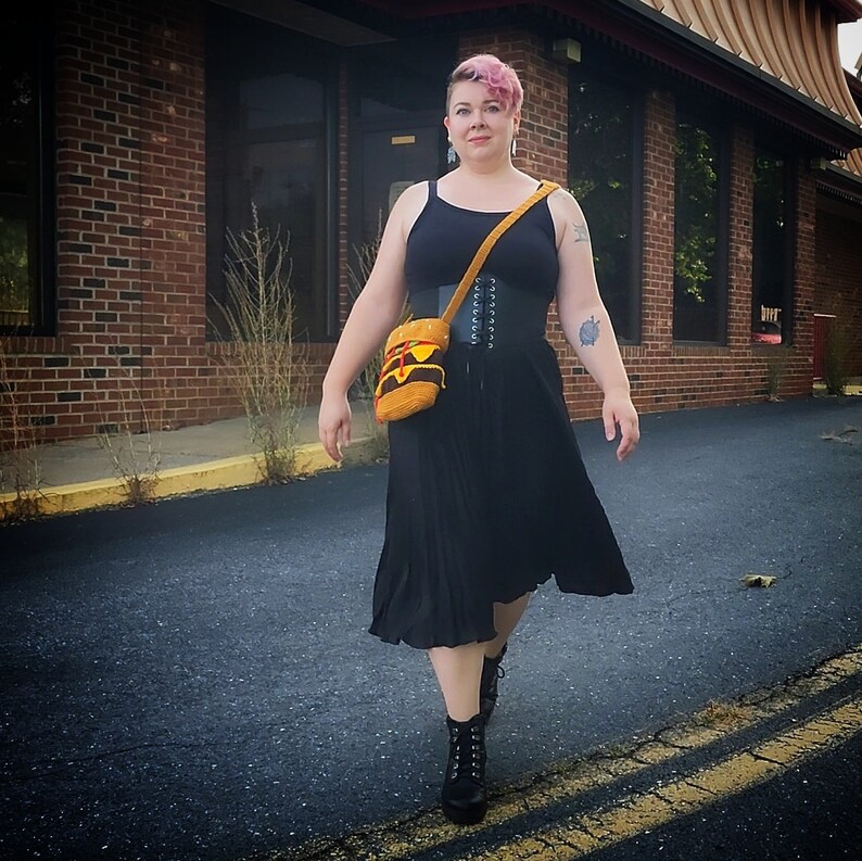 May include: A woman wearing a black tank top, a black corset, and a black pleated skirt. She is carrying a brown and yellow bag with a strap over her shoulder. She is wearing black boots and has pink hair.