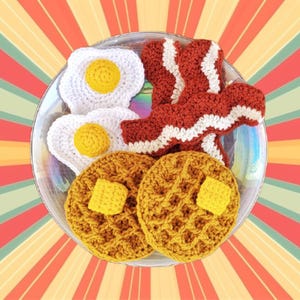 K&ouml;nnte beinhalten: Ein geh&auml;keltes Fr&uuml;hst&uuml;cksset mit zwei gelben Waffeln, zwei wei&szlig;en Eiern und einer Scheibe rot-wei&szlig;em Speck auf einem wei&szlig;en Teller.